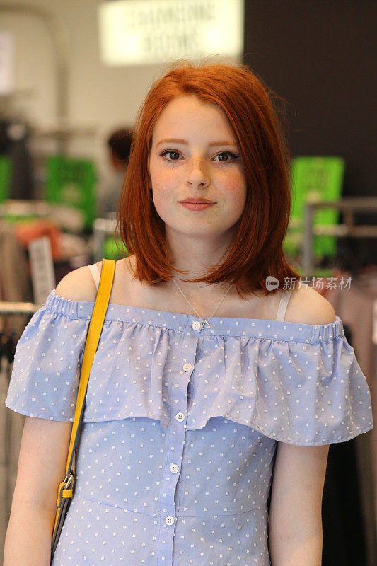 这是一个14 / 15岁的红发少女站在商店里的照片，她穿着一件漂亮的淡蓝色带纽扣的带有白色圆点图案的巴多连衣裙，肩上挎着一个黄色的手提包